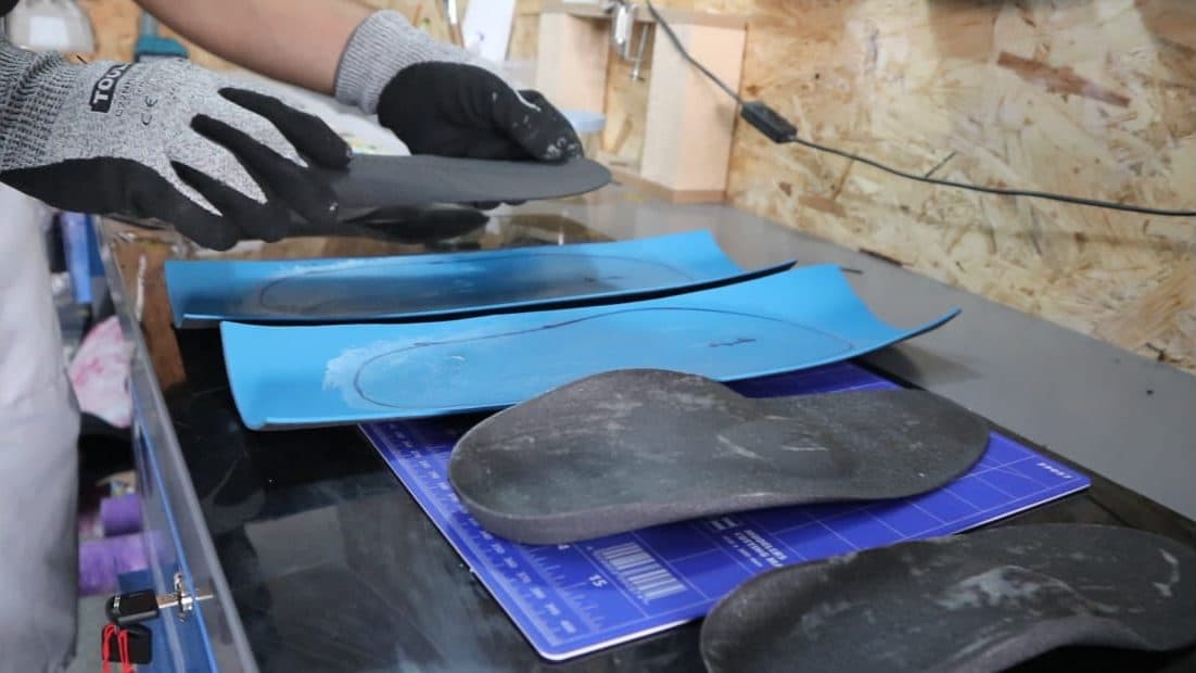 A gloved technician lifting a finished insole from the bench in the BioSoles workshop, with blue moulding sheets and shaped blanks laid out beside it.